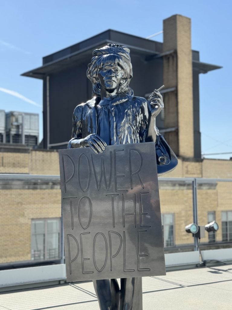 A life-sized aluminum sculpture of a woman smoking a cigarette with a sign that says POWER TO THE PEOPLE