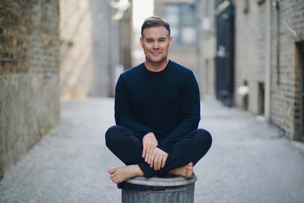 A white, blond-haired man named Ryan Kortman, the founder of Instagram account Whos___who, is dressed in black while sitting cross-legged on a trash can..