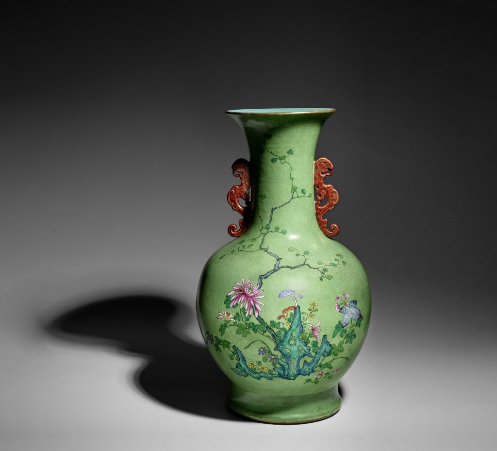 An elaborately decorated green vase with red handles stands against a gray background.