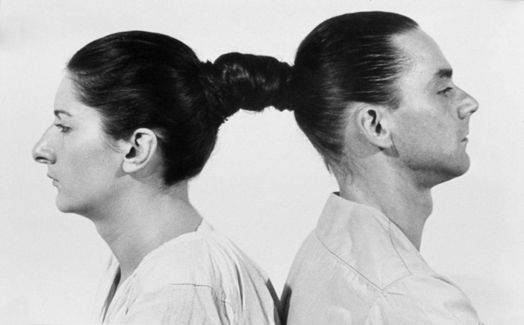 The artists Marina Abramović and Ulay sit back to back with their hair woven together.