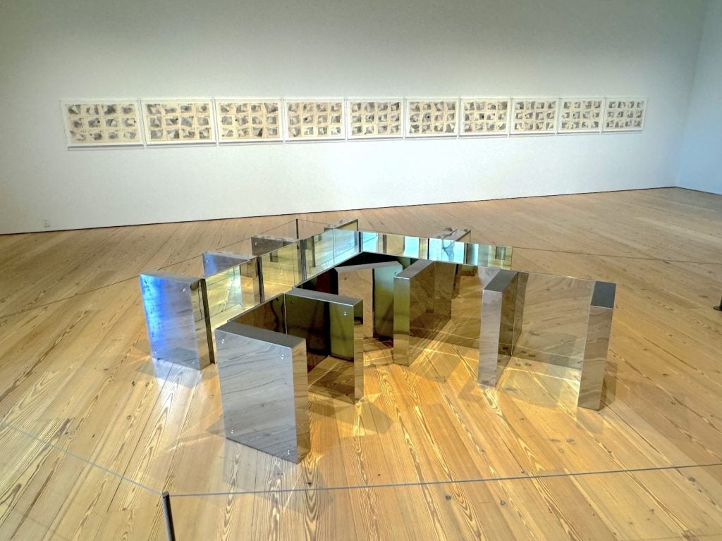 A gallery with metal sculptures on the floor and a series of dense documents on the wall behind