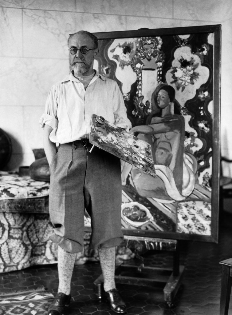 An elderly man, artist Henri Matisse, standing in his studio holding a palette with a painting behind him.