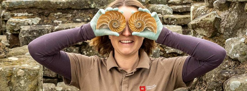 A photograph of a woman from the chest up, holding two halves of a pristine nautilus shell over each eye