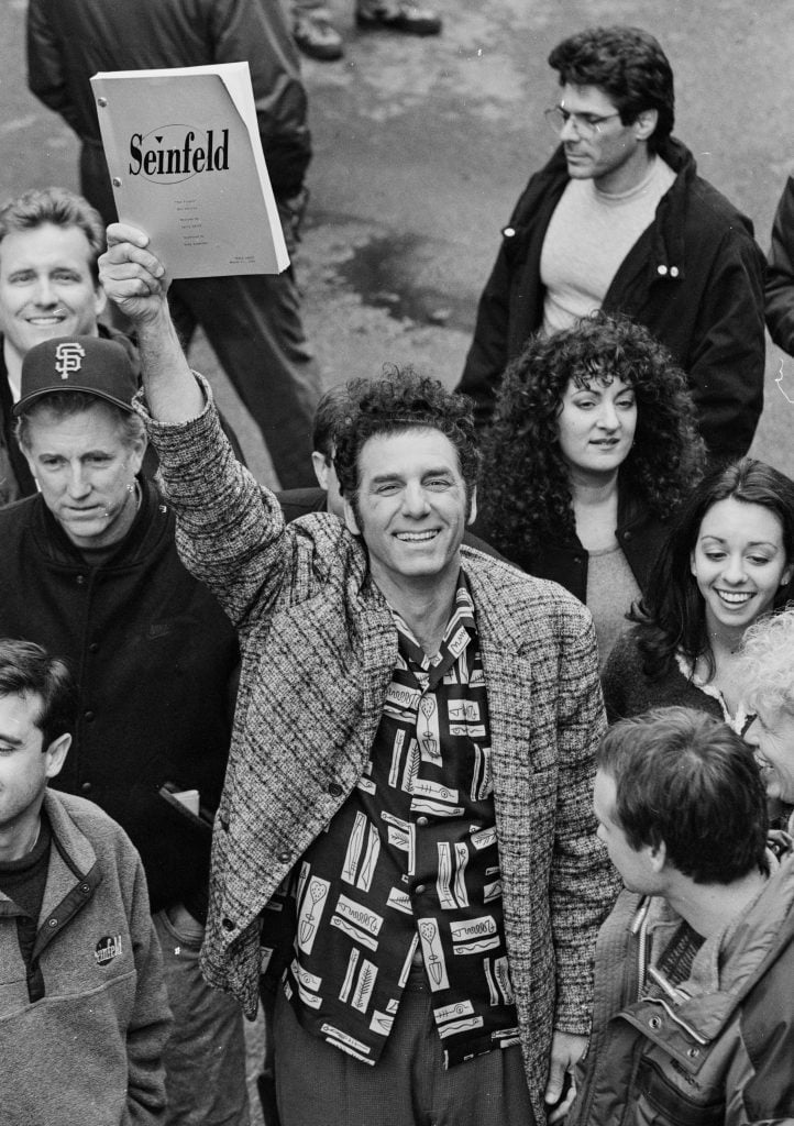 A wild-haired man in a patterned shirt holding up a screenplay with a cover that reads "Seinfeld."