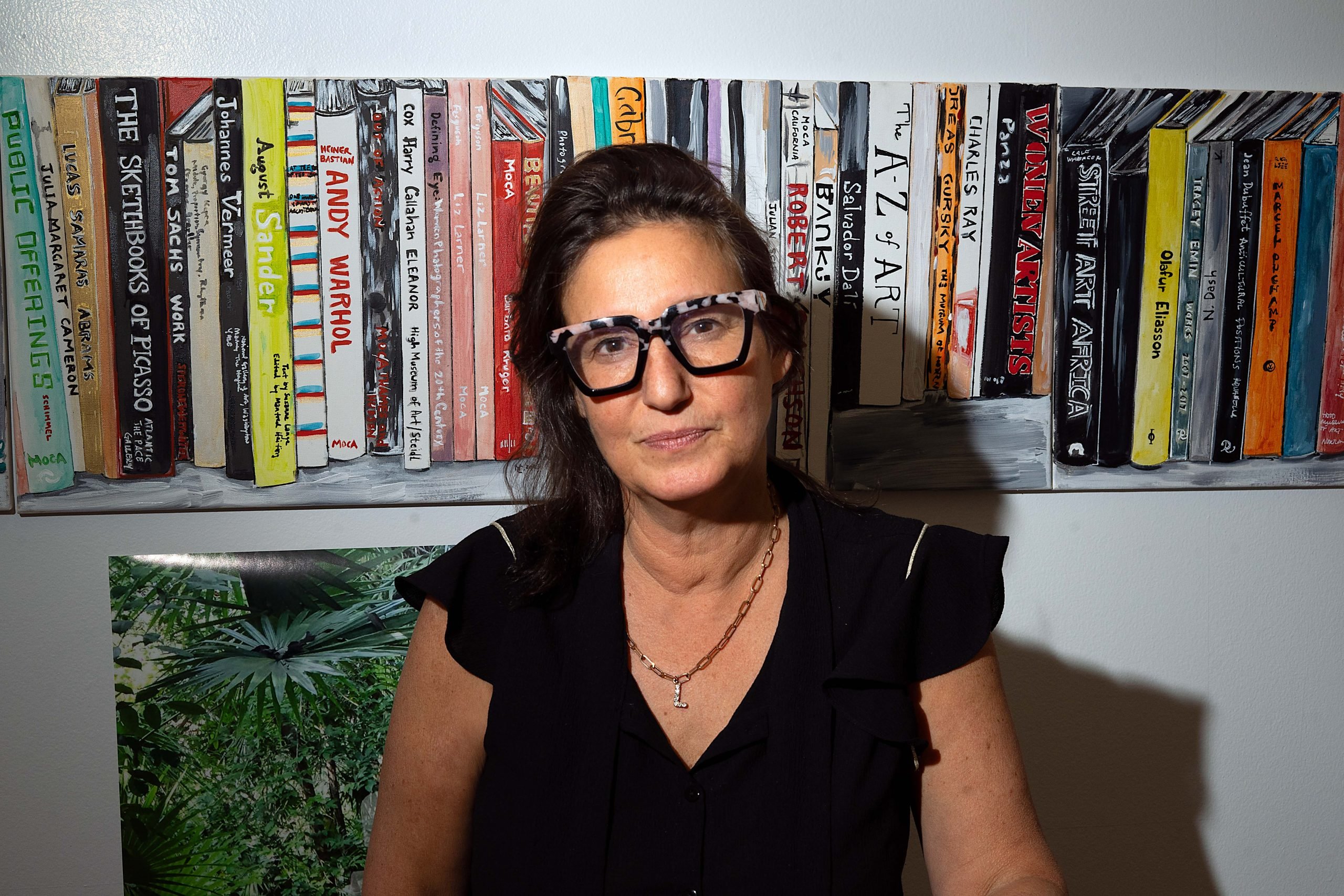 A person stands next to a shelf of books with painted spines that mimic real book titles, suggesting a theme of art and art history as influential factors in their work.