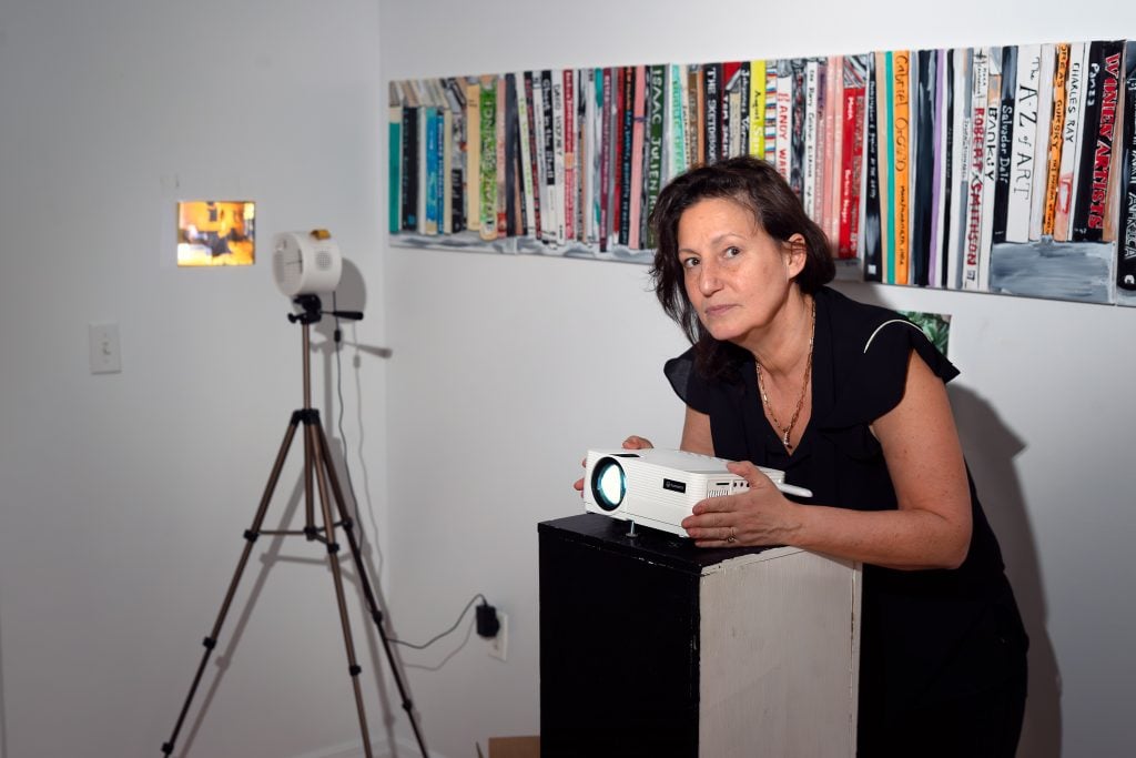 A person posing with a projector, which is used to display images on the wall. A painting of a shelf filled with books, many of which are art-related, is visible in the background.