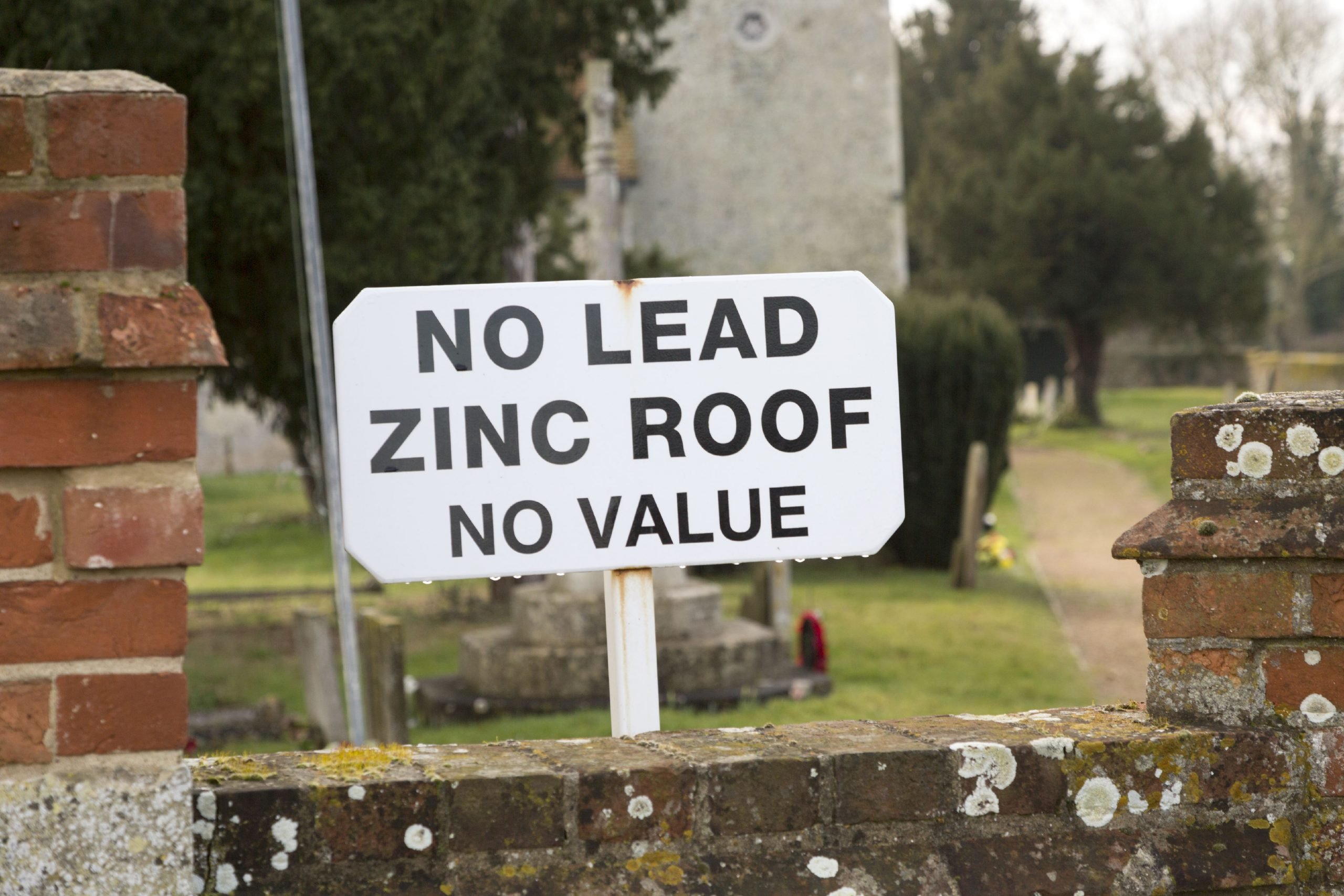 A white sign printed with the words "No Lead. Zine Roof. No Value" planted beside a brick wall to deter theft.