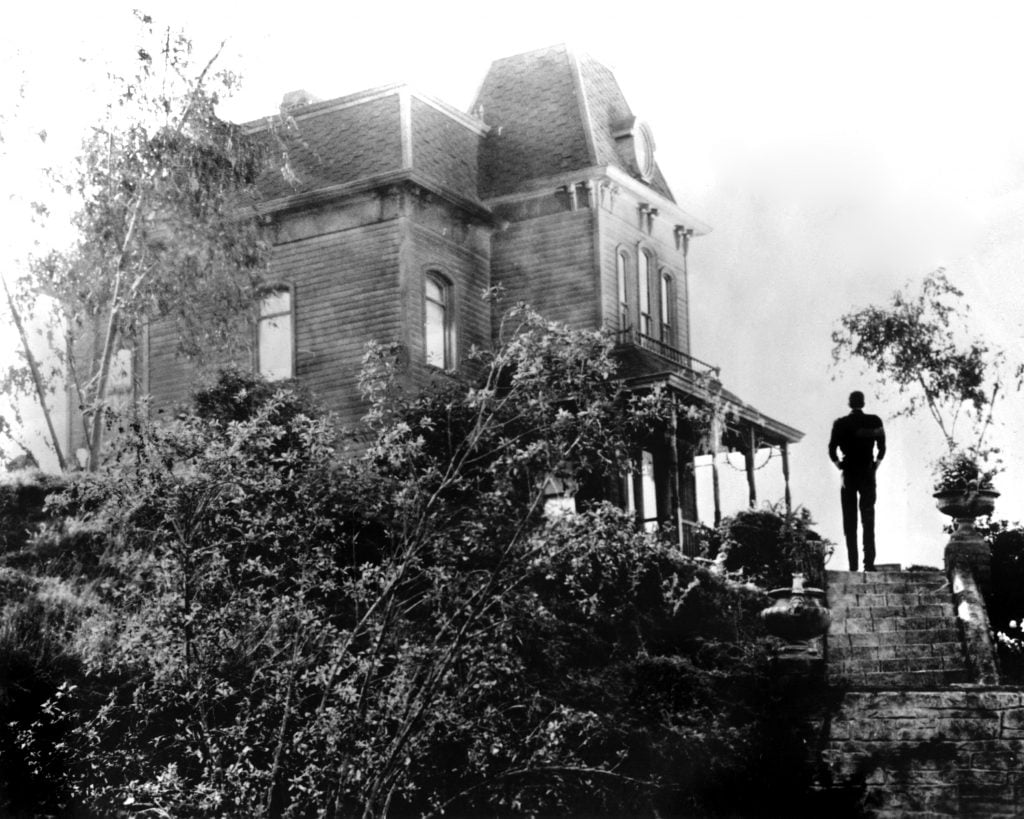 A man stands with his back to the camera in front of a large house.