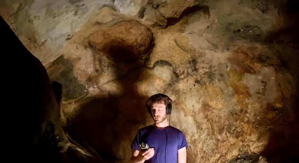 Artist Oliver Beer stands in a Paleolithic cave with a recording device.