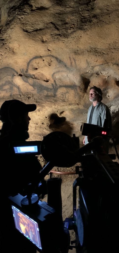 Oliver Beer and Rufus Wainwright with recording equipment in a cave with wall paintings.