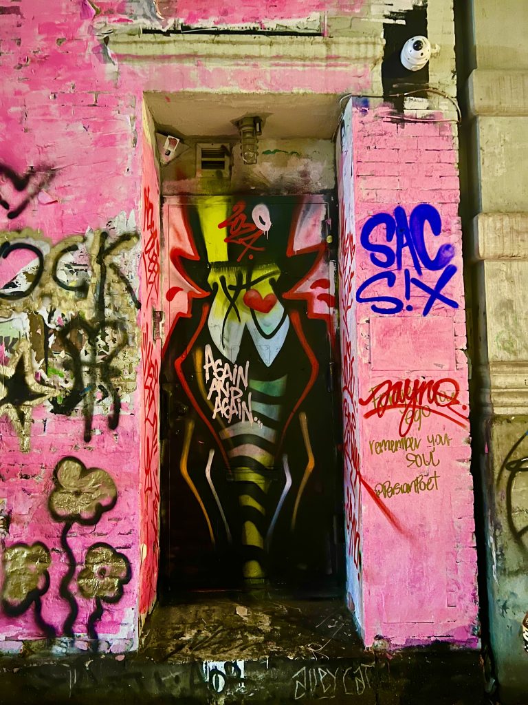 A photograph of red, gold, black and blue graffiti on a pink wall with a spray painted, silhouetted character in a top hot per the style of graffiti writer Optimo