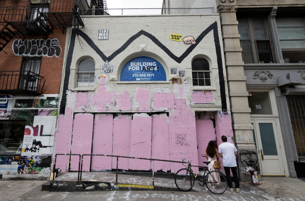A photograph of Basquiat's former studio covered halfway with soft pink paint while two people stand in the lower right and look at it