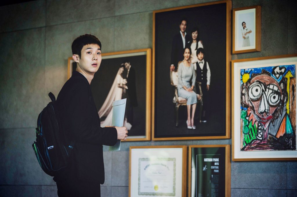 A young man standing beside a wall of framed photos and a childlike drawing.