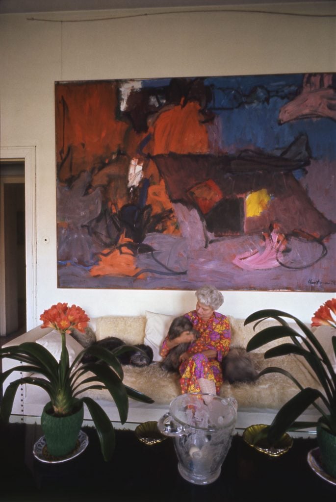 Peggy Guggenheim sits on a plush couch cuddling her dogs. Grace Hartigan's bold abstract work hangs above the couch.
