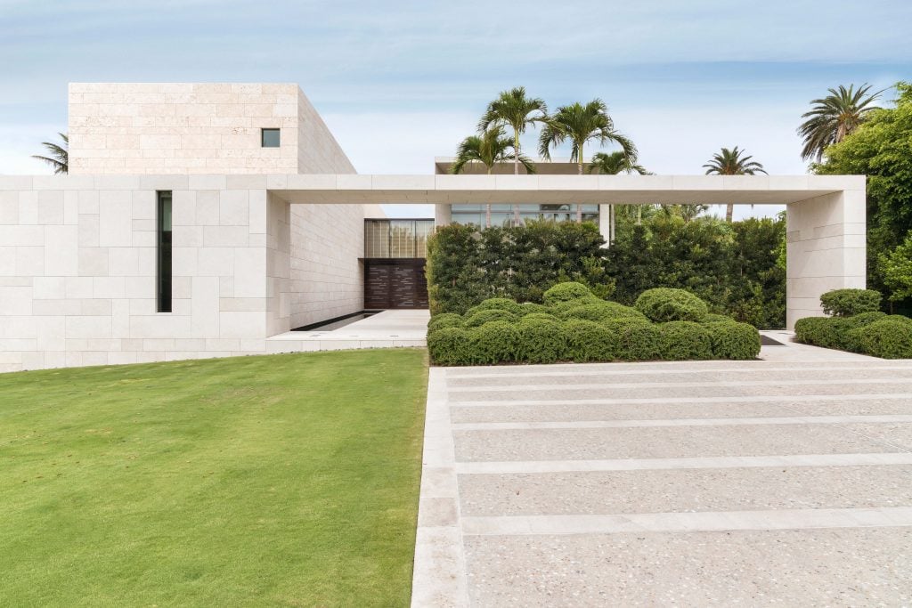 a white modern house has a vast empty lawn in foreground and palm trees behind it 