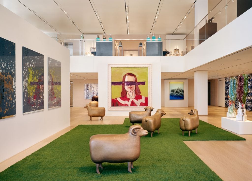 Three bronze sheep sculptures are on top of fake grass in a large gallery space