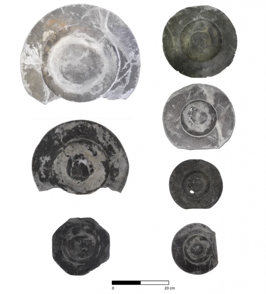 Two colums of seven total pewter plates in a ranger of silver and black hues, each distinctly weathered and chipped from time