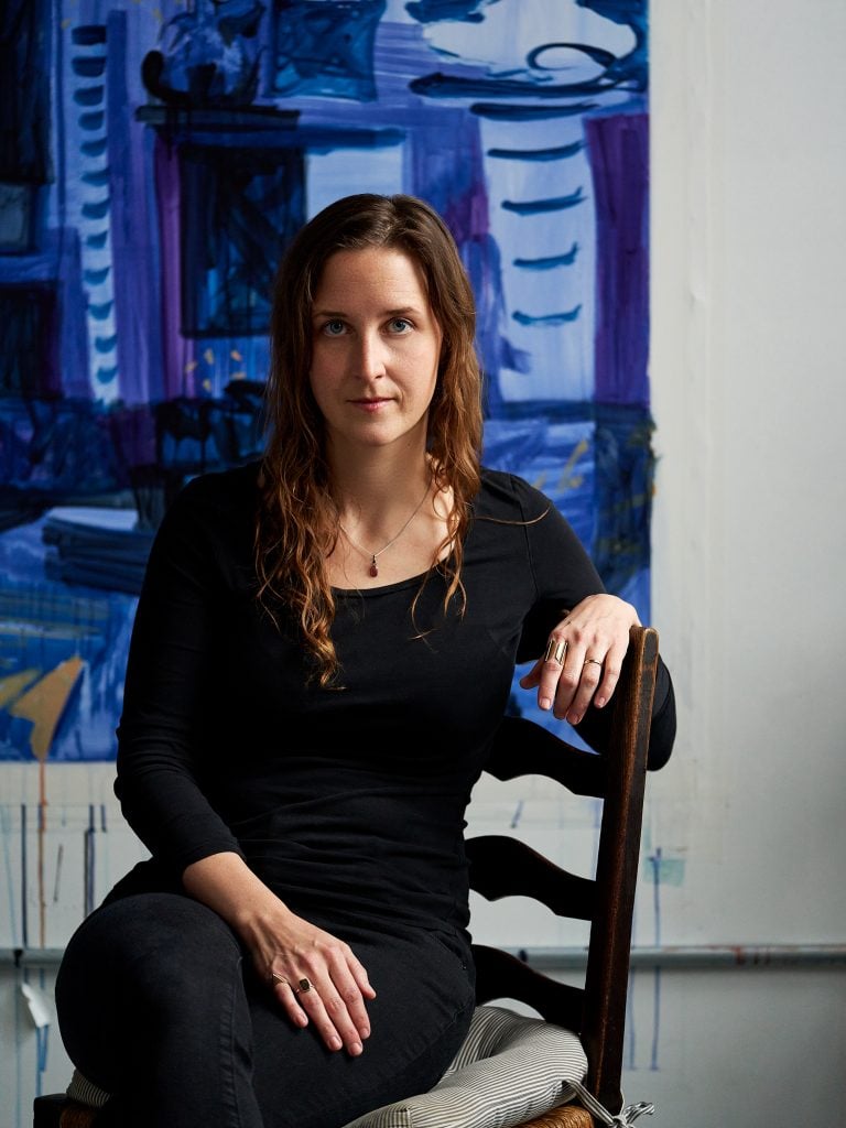 a white woman with long brown hair, wearing black, seated on a chair, a blue painting is visible behind her