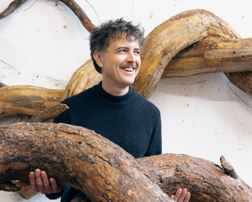 a man is smiling next to a sculpture that looks like the winding trunks of a tree