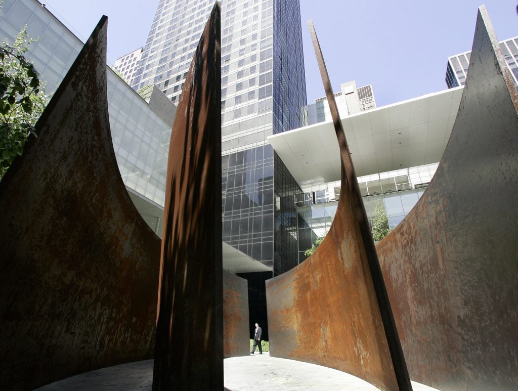 large sheets of rusted metal rise from the ground in an outdoor setting with the skyscrapers of new york visible behnd