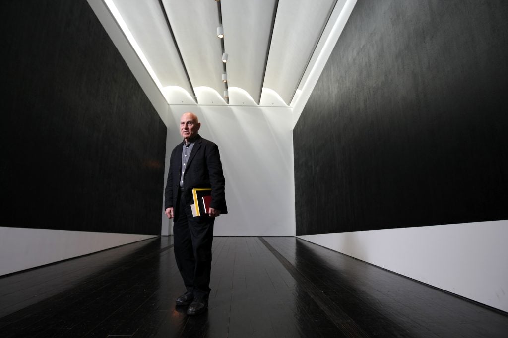 A man in a suit, artist Richard Serra, standing in a gallery hung with two black panels.