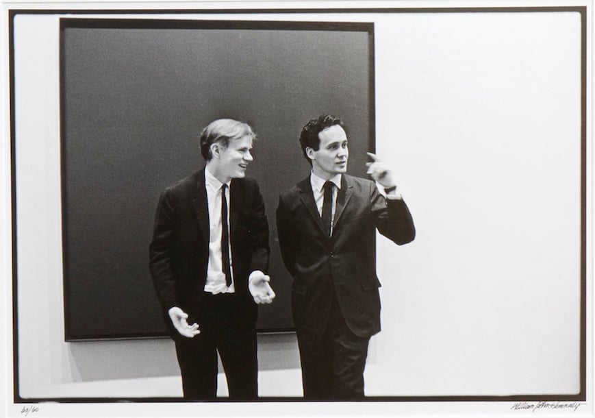 Artists Andy Warhol and Robert Indiana in suits, standing in front of a painting.
