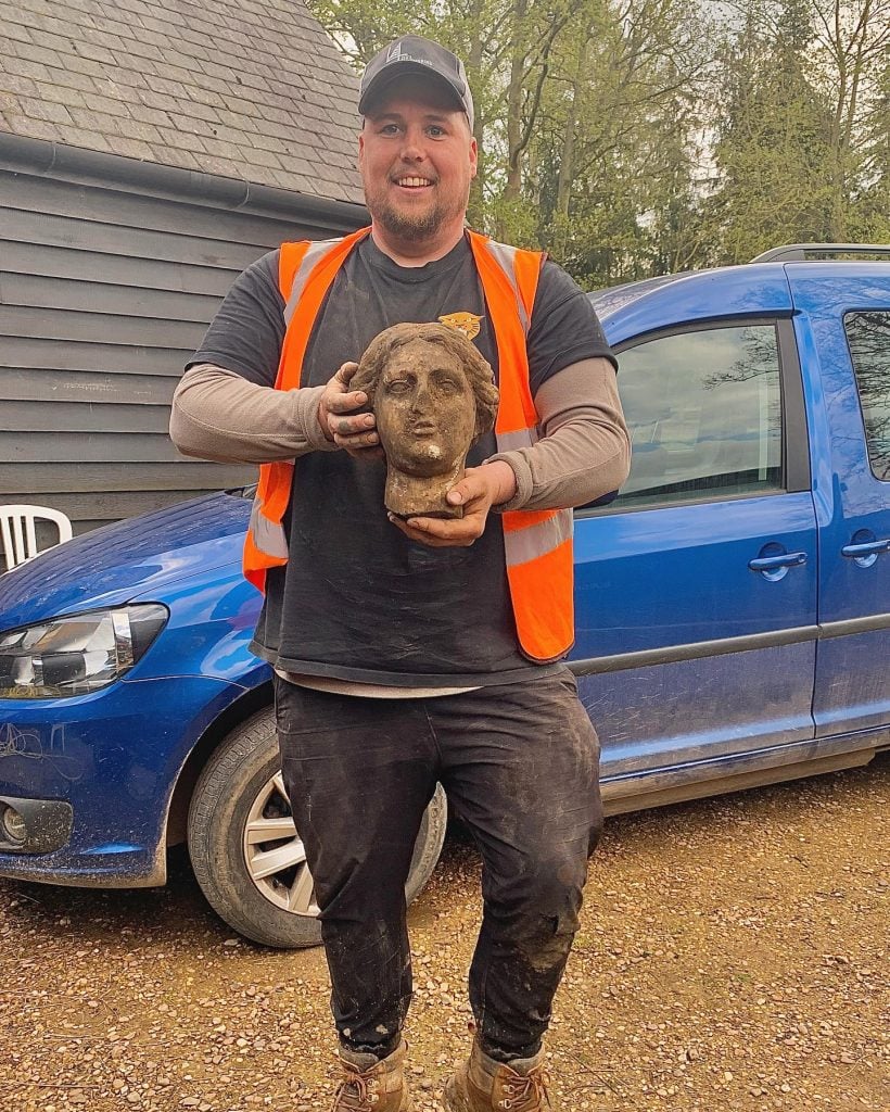 A man in an orange vest holding a Roman bust which is covered in dirt.