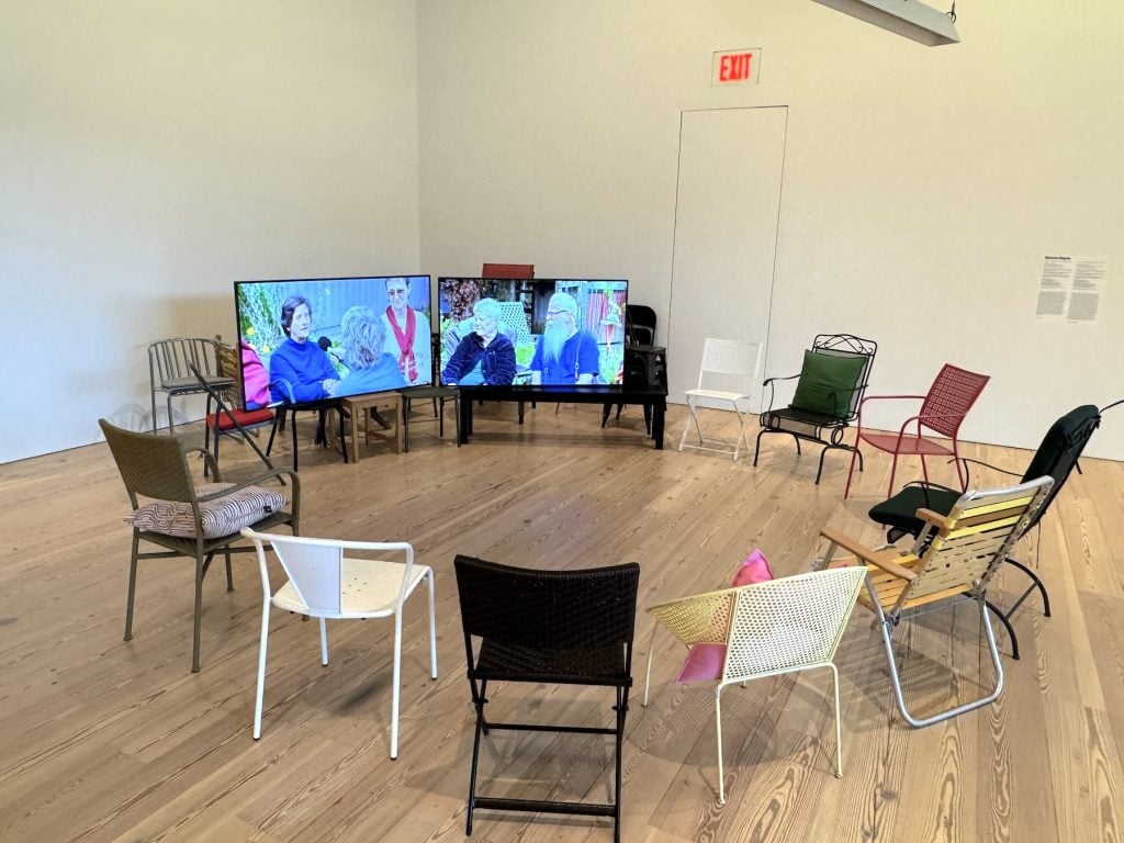Two video monitors featuring people talking surrounded by a circle of chairs