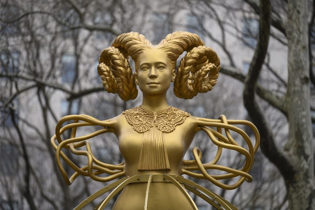 gold statue of a woman with hair braided and coiled like a rams horns. she also has what looks like branches for arms.