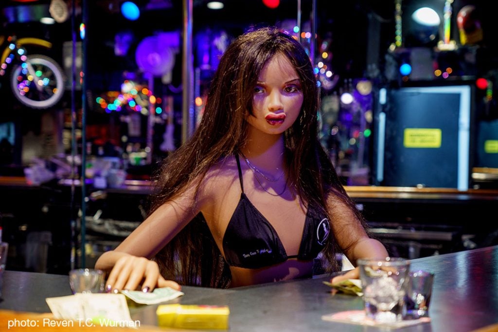 A photograph of an ethnically ambiguous sex doll posed as if working behind a bar