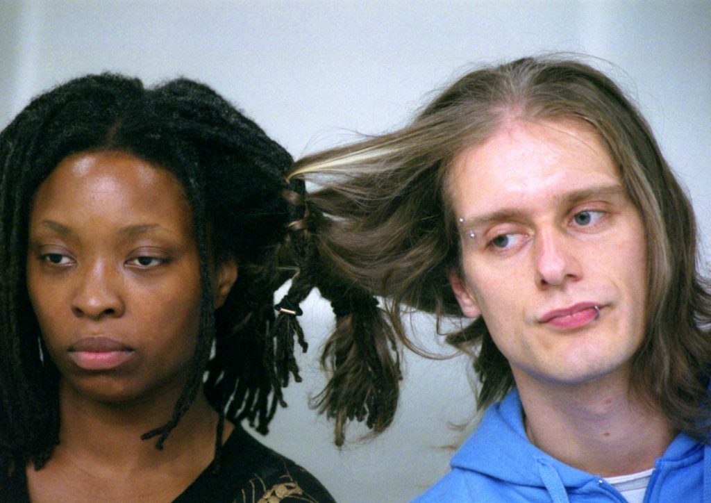 A Black woman and a white man sit side by side, their hair intertwined
