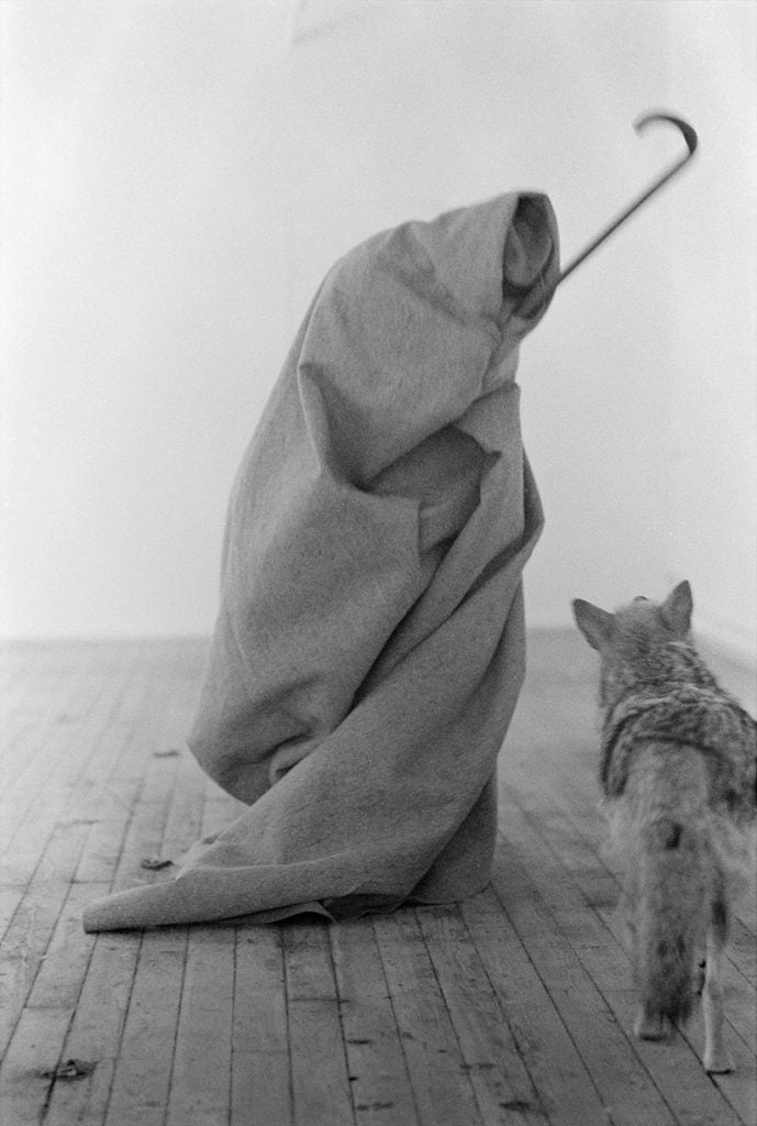 A man wrapped in a felt blanket stands near a coyote, holding out a shepherd's crook.
