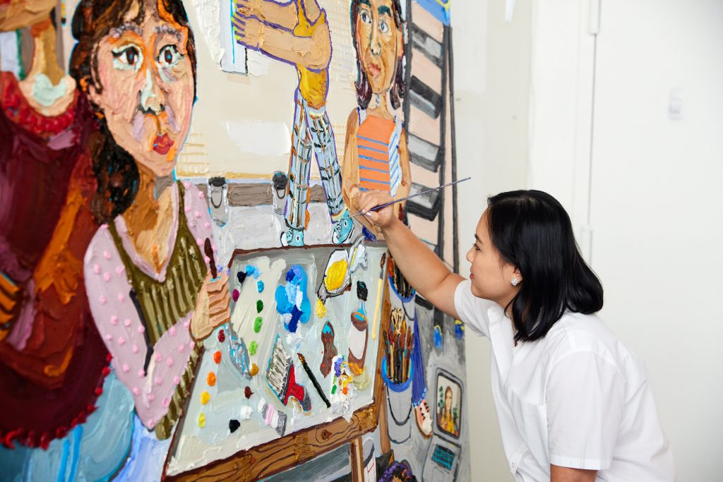 the artist Susan Chen with a paintbrush in hand, working on a painting showing a group of people in an artist studio