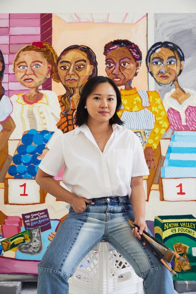 a young asian woman in a white button up shirt and blue jeans, leans in front of a colorful painting