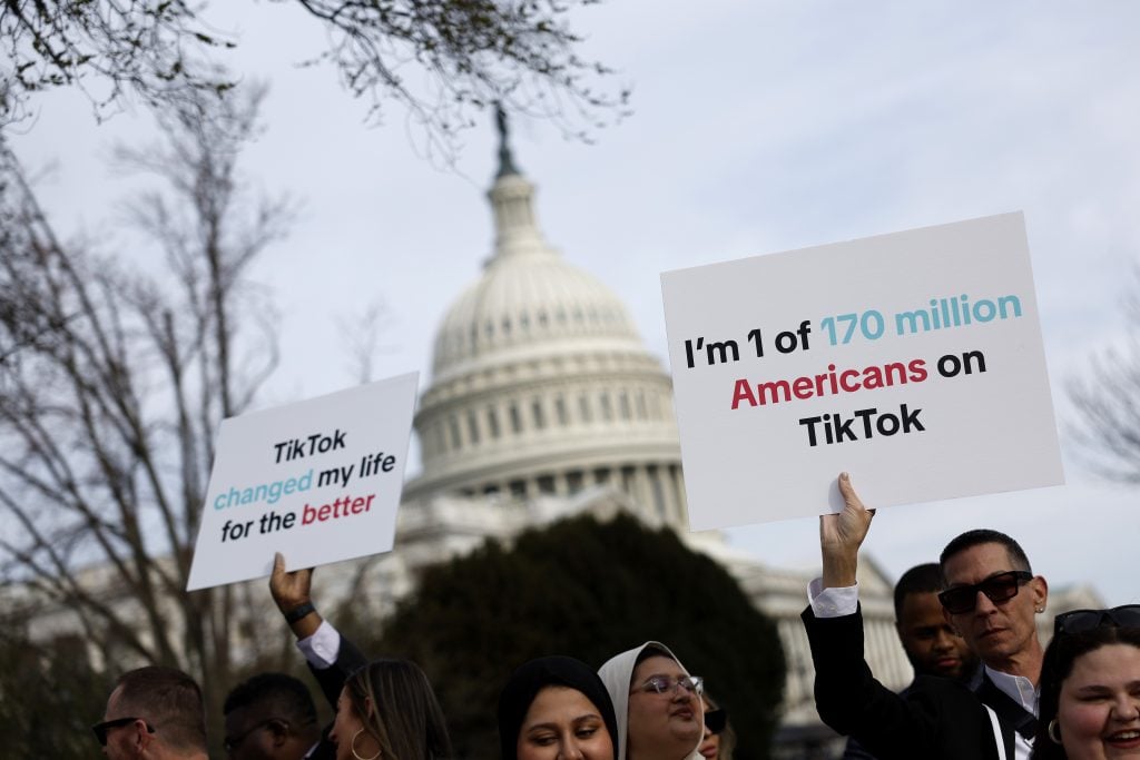The image shows a group of people standing in front of the US Capitol building holding signs. One of the signs reads 