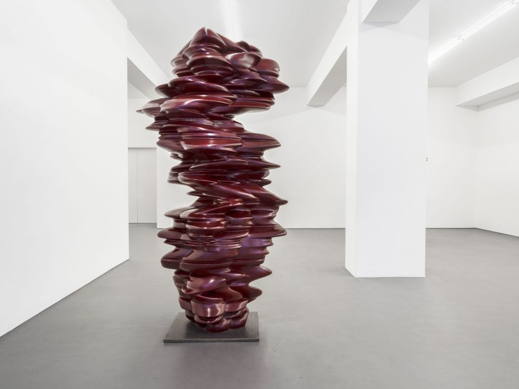 An oxblood red abstract sculpture in a white cube gallery space.