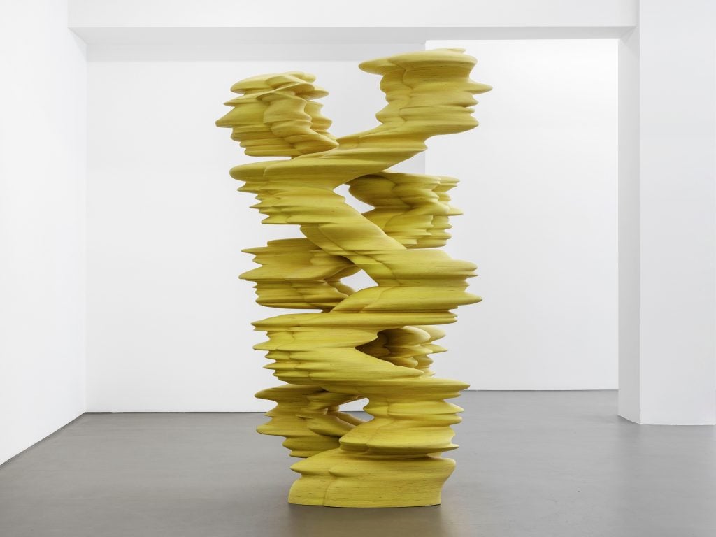 A large-scale yellow abstract sculpture in a white gallery space.