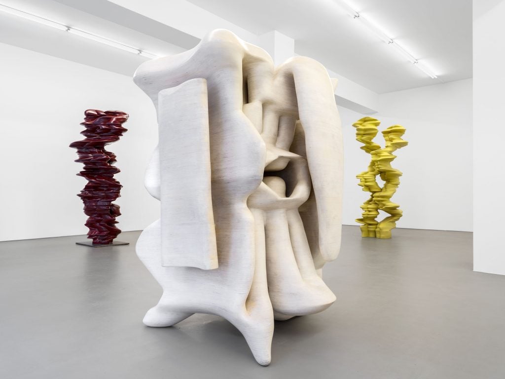 Inside a white gallerie space showing three large-scale abstract sculptures, one oxblood, one yellow, and in the foreground one white.