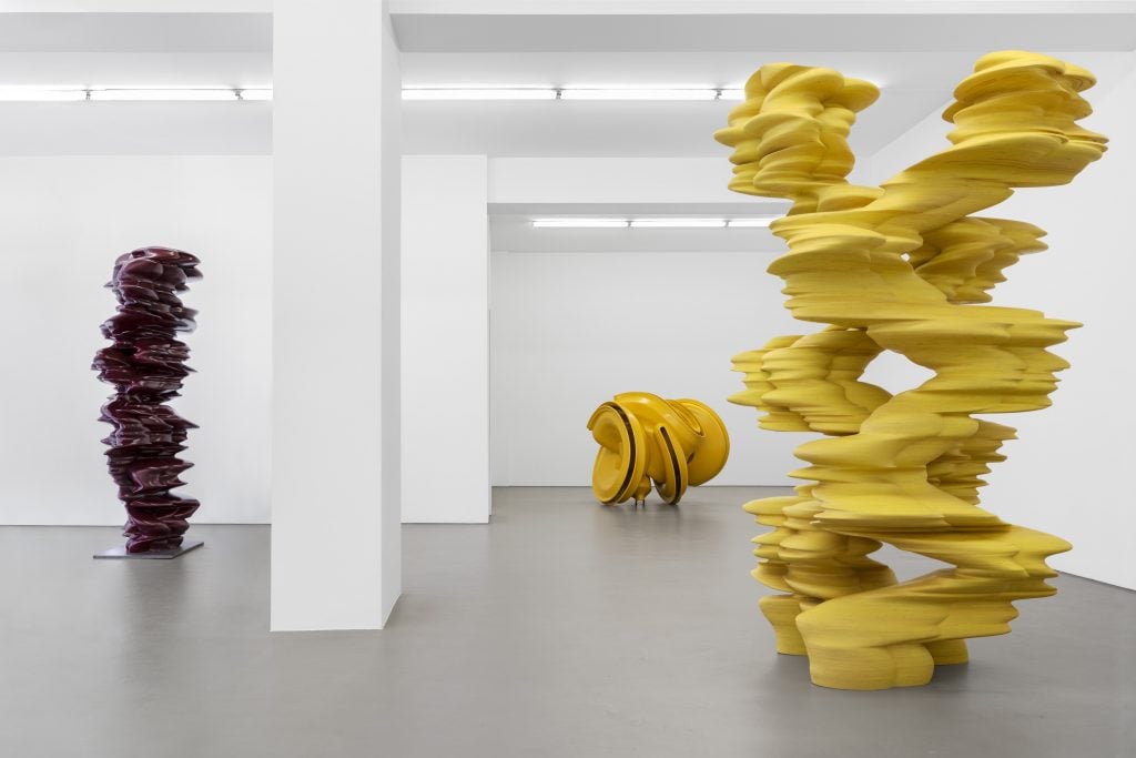 Three large-scale abstract sculptures in a gallery space, two yellow one oxblood.