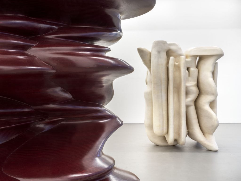 Two abstract sculptures in a white gallery space, in the foreground, an oxblood abstract sculpture is partially cropped out of view. In the background, an off-white abstract sculpture.