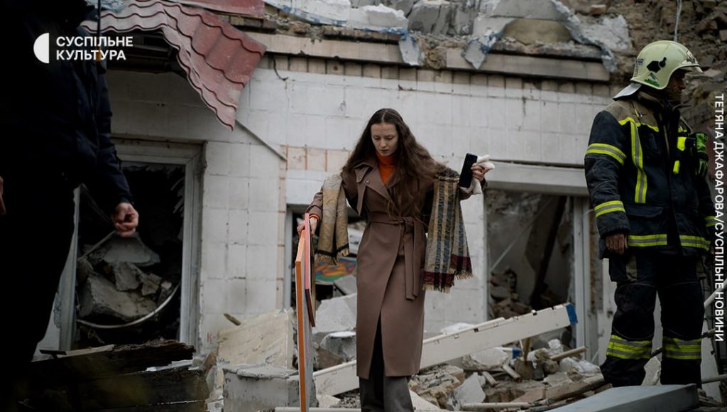 A woman in a tan coat and orange scarf exits a bombed building with a crutch in one hand and a phone in the other. She is accompanied by a firefighter in full gear standing amidst the rubble