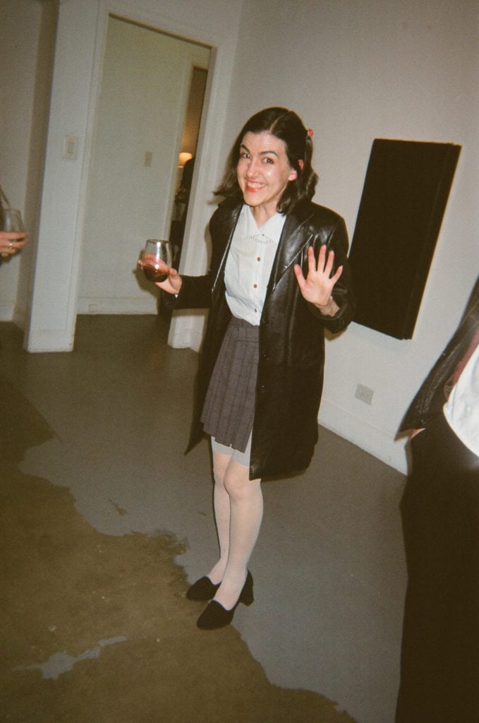 A woman in a pleated skirt, leather jacket, and heels, holding a glass of red wine smiles for the camera.