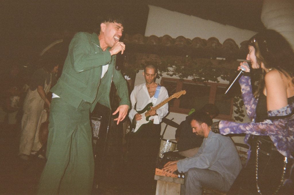 a man singing into a microphone wears a matching green pantsuit, a woman in a flowy purple outfit sings across from him and a person in a white shirt and black pants plays the guitar in the background.