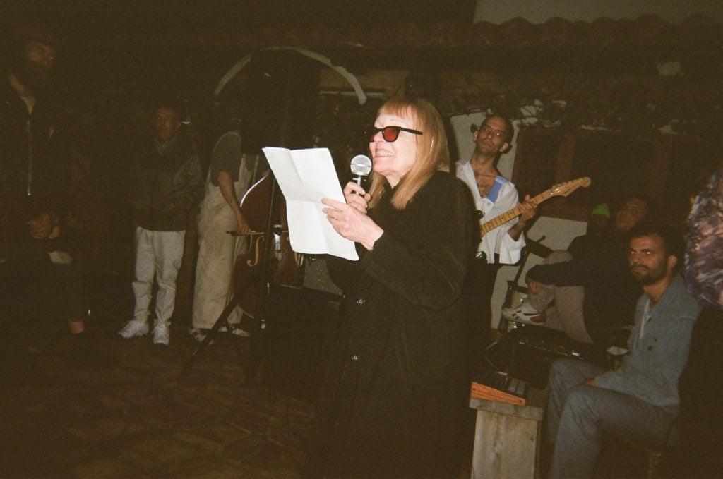 A woman in all black and sunglasses reads a poem into a microphone in a dimly lit room.