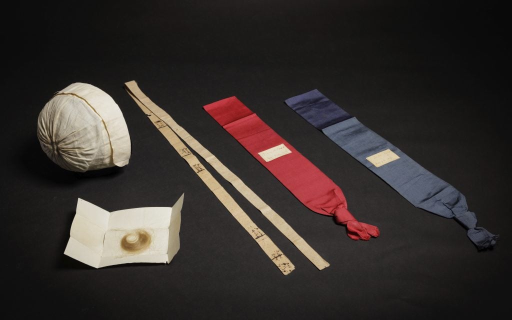 A photograph of five objects, including an envelope with hair in it, a bonnet, two beige measuring sticks, and two sashas—one red and one blue