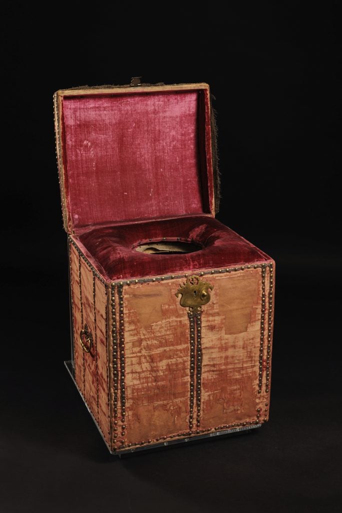 A photograph of a 17 century close stool (toilet) which appears as a wooden box open on top to reveal red velvet interior and a hole