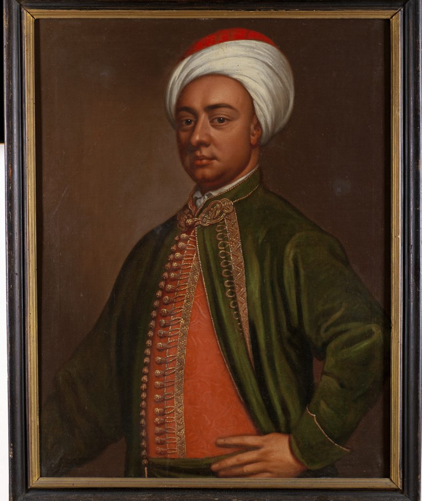 A photograph of a framed portrait of a tanned man wearing a green coat, red vest, and white turban