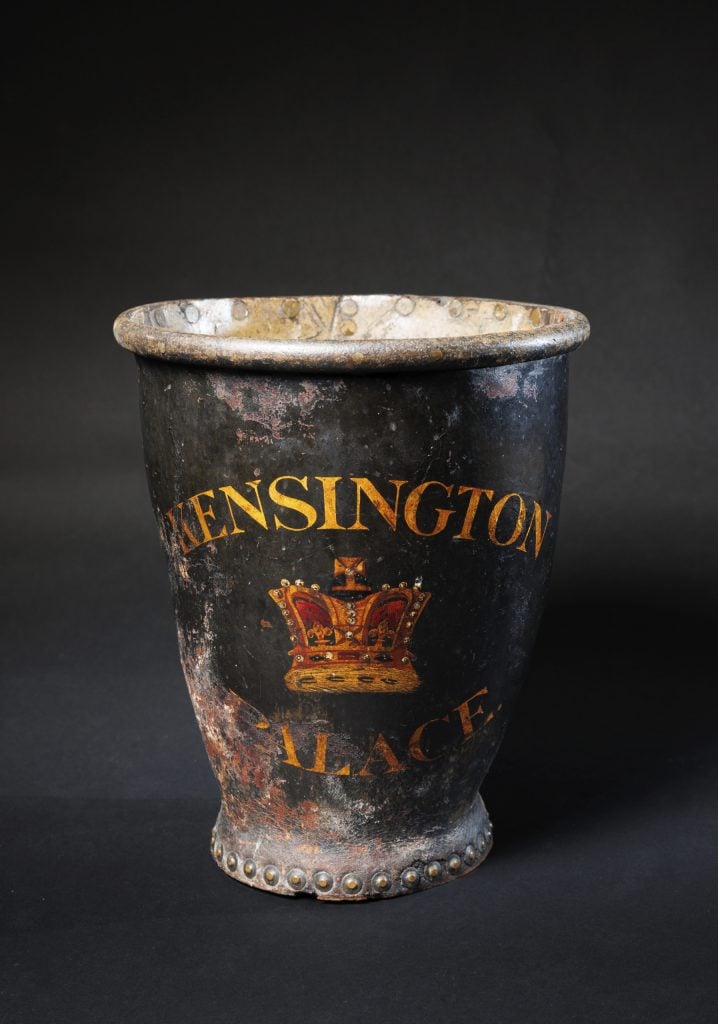 Photograph of a worn black leather fire bucket embalzoned in bronze with 'Kensington Palace' and a crown between those two words
