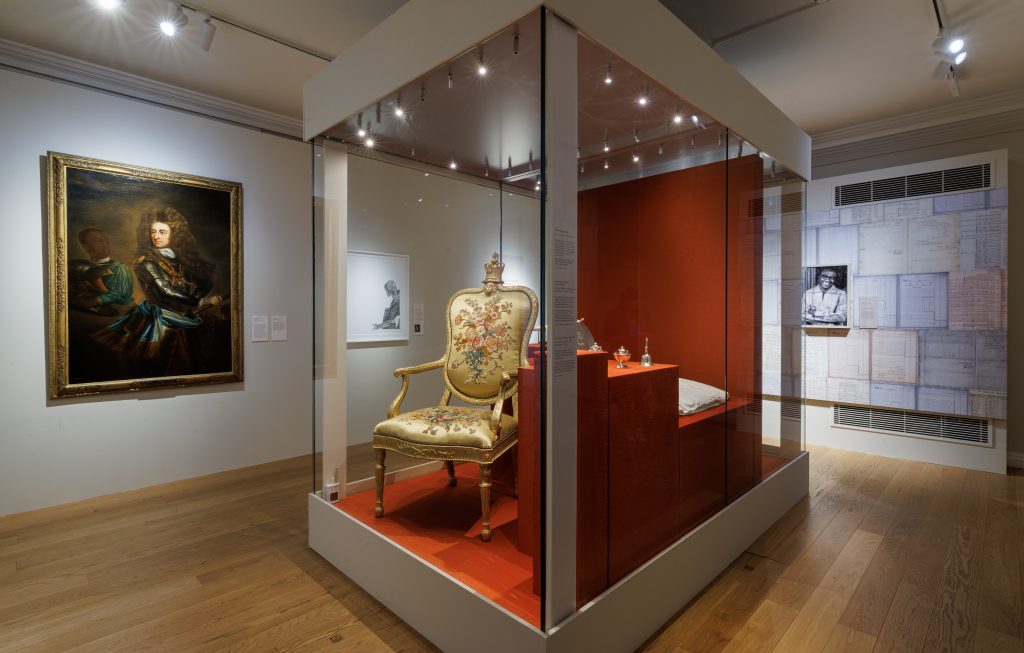 A photograph of a room in the 'Untold Lives' exhibit at Kensington Palace depicting an ornate chair and other finery in a glass case surrounded by paintings and installations of records