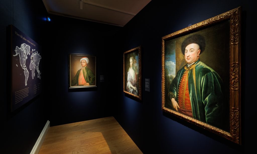 A photograph of a room in the museum exhibit 'Untold Lives' at Kensington Palace featuring three 18th century painted portraits on a navy blue wall